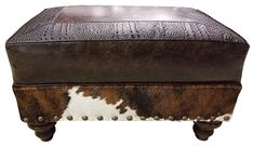 a brown and white cowhide ottoman with metal studs on the legs, sitting against a white background