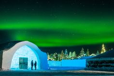 two people standing in front of an igloose under the northern sky with green lights