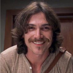 a man with long hair and a moustache smiles at the camera in front of a door