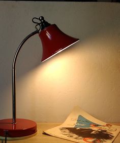 a red desk lamp sitting on top of a wooden table next to a book and magazine