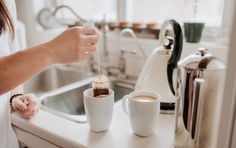 a woman pours coffee into two mugs on the counter in her home kitchen