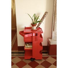 a red table with a plant on top and some books in the bottom shelf next to it