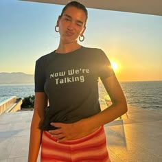 a woman standing in front of the ocean with her hands on her hips, wearing a t - shirt that says now we're talking