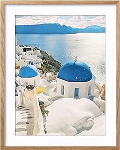 a framed photograph of some white and blue buildings by the water with mountains in the background