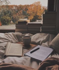 an open book, coffee cup and pen on top of a bed in front of a window