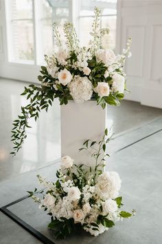 White wooden pillars hold large floral arrangements for a wedding ceremony, with white hydrangeas, white roses, and larkspur, to enhance the class white wedding Floral Urns Wedding Ceremony, Pew Florals Wedding, Wedding Altar Arrangements, Outside Wedding Ceremony Flowers, Staircase Floral Arrangement, Floor Arrangements Wedding Ceremony, Plinth Wedding Flower Arrangements, Church Wedding Decorations Alter Floral Arrangements, Plinth Flowers Wedding