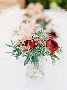 red and white flowers in a clear vase on a table with greenery around it