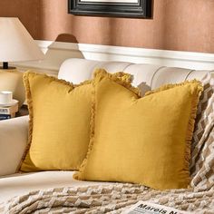 two yellow pillows sitting on top of a white couch next to a book and lamp