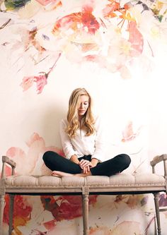 a woman sitting on top of a bench in front of a wall with flowers painted on it