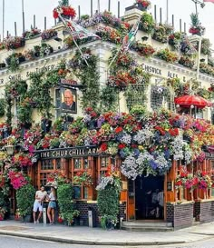 a building covered in lots of flowers and hanging plants on it's side with people standing outside