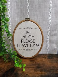 a cross stitch hoop with the words live laugh, please leave by on it next to some greenery