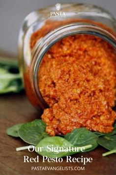 a jar filled with red pesto next to green leaves on top of a wooden table