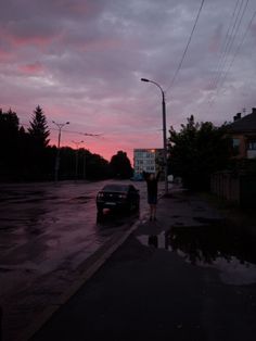 a person standing next to a car on a wet street at dusk with the sun setting