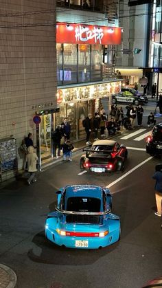 a blue car driving down a street next to tall buildings with people standing on the sidewalk