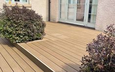 a wooden deck with plants and bushes in front of the house on a sunny day