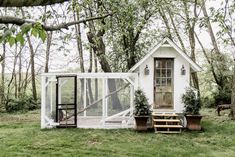 a small white chicken coop in the middle of a field with potted plants next to it