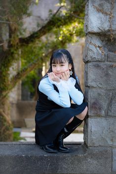 a young woman sitting on the ground next to a stone wall with her hand under her chin