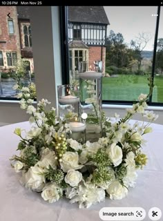 a centerpiece with white flowers and greenery sits on a table in front of a large window