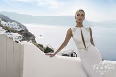 a woman in a white dress leaning against a wall near the ocean and buildings on the other side