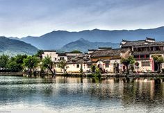 an old chinese village on the edge of a body of water with mountains in the background