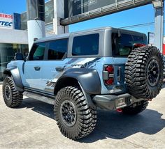 a large gray truck parked in front of a dealership with huge tires on it