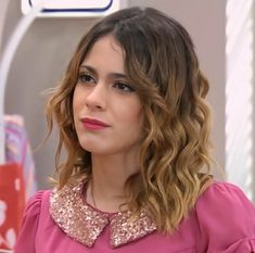 a close up of a person wearing a pink shirt and gold collared top with long curly hair