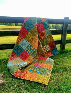 a patchwork quilt sitting on the grass next to a fence in front of a field