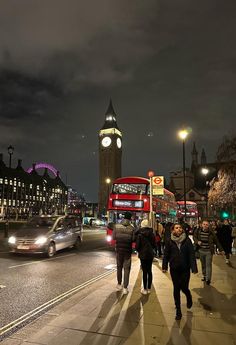 people walking on the sidewalk in front of a clock tower at night with cars passing by