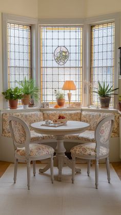 a corner table and chairs in front of two windows with potted plants on them