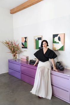 a woman in a black top and white skirt leaning on a dresser with purple drawers