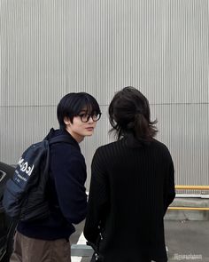 two people standing next to each other in front of a building with a black car