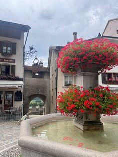 a fountain with red flowers on it in the middle of a town square surrounded by buildings