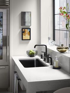 a kitchen sink sitting under a window next to a vase with flowers and fruit on it