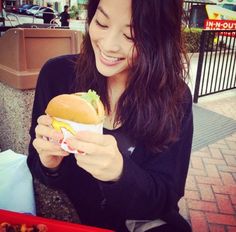 a woman holding a sandwich in her right hand and smiling at the camera while sitting on a bench