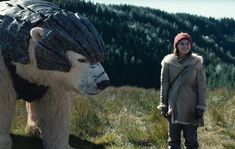 a woman standing next to a giant polar bear in a field with mountains behind her