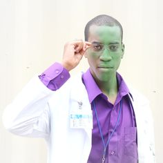 a man with green paint on his face wearing glasses and a purple shirt is standing in front of a white wall