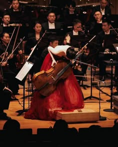 a woman in a long red dress is playing the violin and orchestra behind her on stage