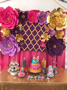 a table topped with lots of cakes and desserts next to a wall covered in paper flowers