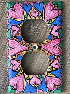 a colorful light switch cover with hearts painted on the front and back of it, sitting on a wooden surface