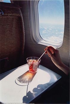 an airplane seat with a drink on the table next to it and another person's hand holding a straw