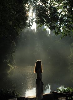 a woman standing in front of a body of water with the sun shining through trees