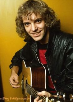 a man with curly hair holding a guitar and smiling at the camera while wearing a black leather jacket