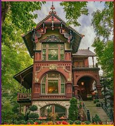 an old victorian style house in the middle of some trees and bushes with flowers around it