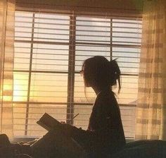 a woman sitting in front of a window writing on a piece of paper while the sun is setting