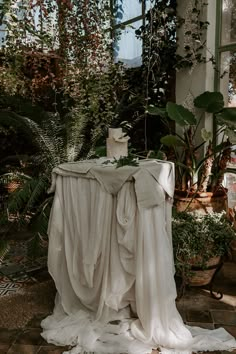 the table is covered with white cloths and has a cake on it, surrounded by greenery