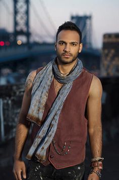 a man standing in front of a bridge wearing a brown vest and scarf with his hand on his hip