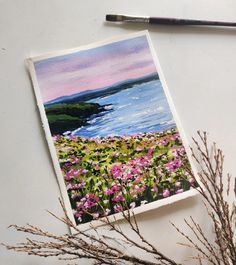 a painting is shown next to some flowers and a paintbrush on the table with water in the background