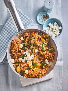 a pan filled with rice and vegetables on top of a blue table cloth next to two spoons