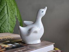 a white bird figurine sitting on top of a stack of books next to a green plant