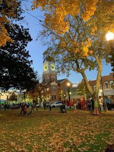 many people are walking around in the grass near trees with yellow leaves on them and one person is holding a cell phone